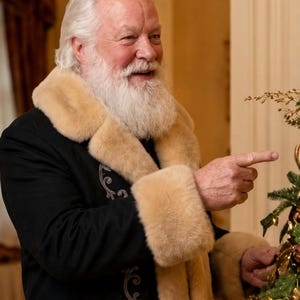 May include: A person with white hair and a full white beard wearing a black coat with a thick beige faux fur collar and cuffs. The coat features intricate silver embroidery along the front opening. The person is pointing toward a decorated tree.