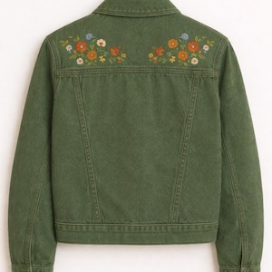 May include: An olive green denim jacket featuring a classic collar, long sleeves with buttoned cuffs, and two symmetrical floral embroidery patterns in orange, yellow, and blue thread across the upper back panel, hanging on a black hanger.