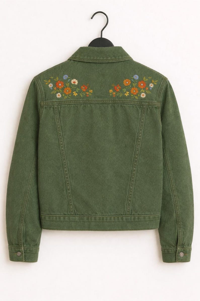 May include: An olive green denim jacket featuring a classic collar, long sleeves with buttoned cuffs, and two symmetrical floral embroidery patterns in orange, yellow, and blue thread across the upper back panel, hanging on a black hanger.