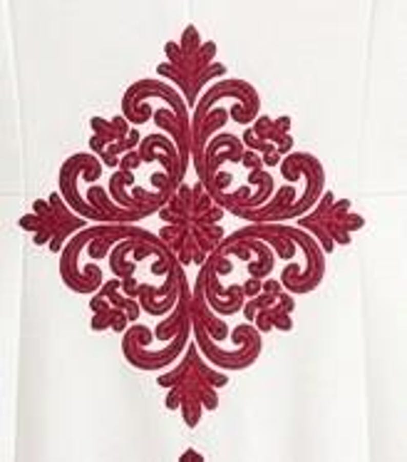 May include: A white fabric surface featuring a large, symmetrical, diamond-shaped embroidery pattern in deep red thread. The design consists of intricate, swirling scrollwork and floral-inspired motifs centered in a vertical orientation.