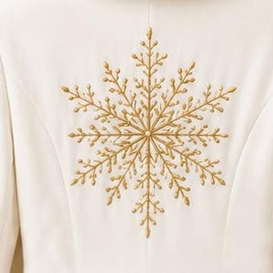 May include: A white garment featuring a large, centered, gold-colored embroidered snowflake design. The embroidery consists of intricate, branching lines radiating from a central point, creating a detailed, symmetrical, and metallic-toned pattern.
