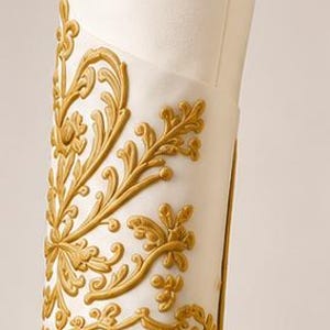 May include: A white fabric sleeve cuff featuring raised gold embroidery in a scrolling floral pattern. The embroidery covers the outer surface of the cuff, which is finished with a thin gold border along the bottom edge of the garment sleeve.