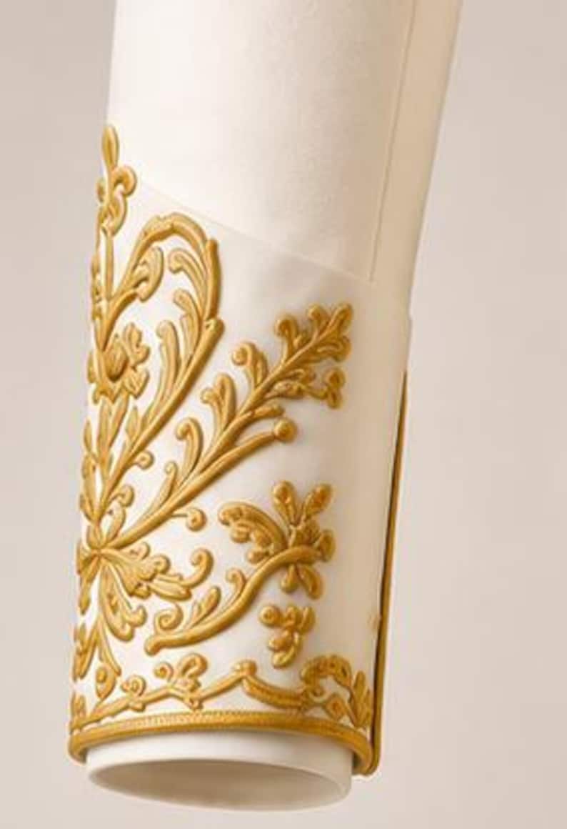 May include: A white fabric sleeve cuff featuring raised gold embroidery in a scrolling floral pattern. The embroidery covers the outer surface of the cuff, which is finished with a thin gold border along the bottom edge of the garment sleeve.
