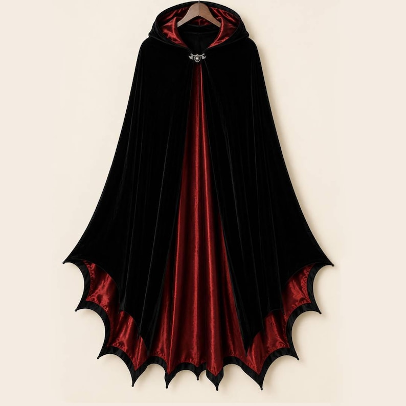 May include: A floor-length hooded cloak featuring a black velvet exterior and a deep red satin lining. The hem is cut into a distinct scalloped bat-wing shape. A silver-toned metal clasp with a central circular detail secures the garment at the neck.