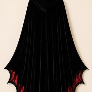 May include: A floor-length black velvet hooded cloak featuring a deep red satin lining. The hem of the garment is cut into a distinct bat-wing or scalloped shape, creating pointed edges along the bottom border of the cape.