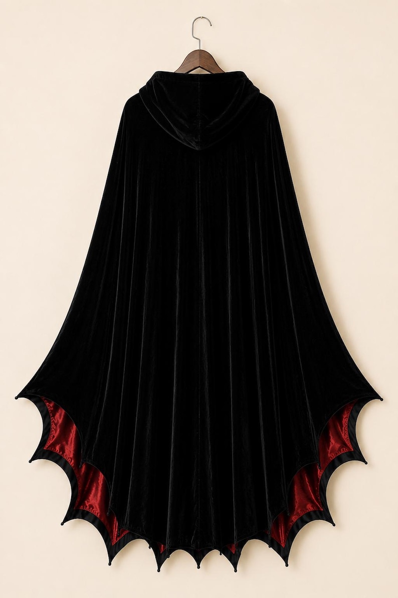 May include: A floor-length black velvet hooded cloak featuring a deep red satin lining. The hem of the garment is cut into a distinct bat-wing or scalloped shape, creating pointed edges along the bottom border of the cape.