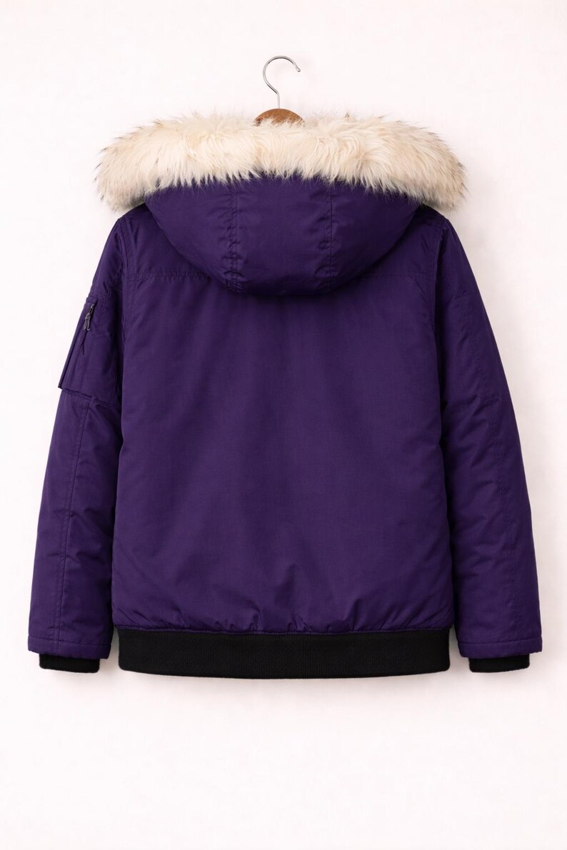 May include: A purple bomber jacket with a hood and a faux fur trim. The jacket has a black ribbed hem and cuffs. A small pocket with a zipper is on the left sleeve. The jacket is hanging on a wooden hanger.