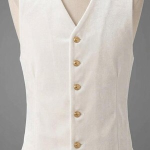 May include: A white velvet vest featuring a V-neckline and five gold-toned buttons down the front. The garment is sleeveless with a pointed hemline, showcasing a smooth, soft-textured fabric finish on a mannequin torso.