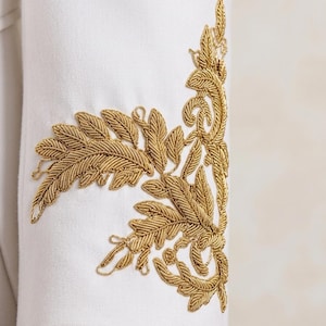 May include: A white long-sleeved garment featuring intricate gold metallic thread embroidery on the cuff. The embroidery depicts a detailed botanical leaf and vine pattern that extends vertically along the side of the sleeve.