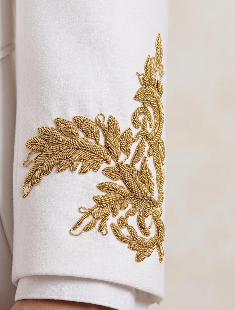 May include: A white long-sleeved garment featuring intricate gold metallic thread embroidery on the cuff. The embroidery depicts a detailed botanical leaf and vine pattern that extends vertically along the side of the sleeve.