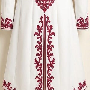 May include: A white long coat featuring deep red embroidery in a symmetrical floral and scroll pattern. The design is placed along the hem, the cuffs, and vertically down the center back, with a prominent medallion motif at the upper back.