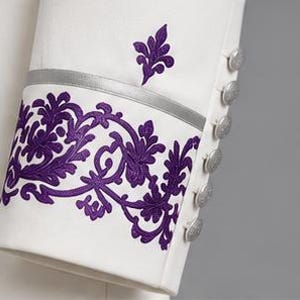 May include: A white sleeve featuring a vibrant purple floral embroidery pattern along the cuff, accented by a thin horizontal silver band. A row of six round silver buttons is aligned vertically along the side of the sleeve cuff.