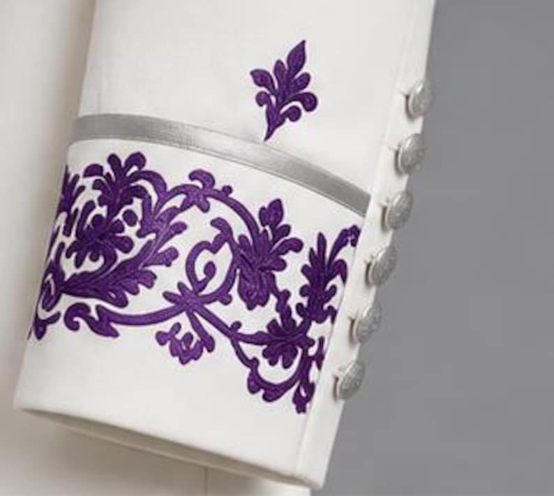 May include: A white sleeve featuring a vibrant purple floral embroidery pattern along the cuff, accented by a thin horizontal silver band. A row of six round silver buttons is aligned vertically along the side of the sleeve cuff.