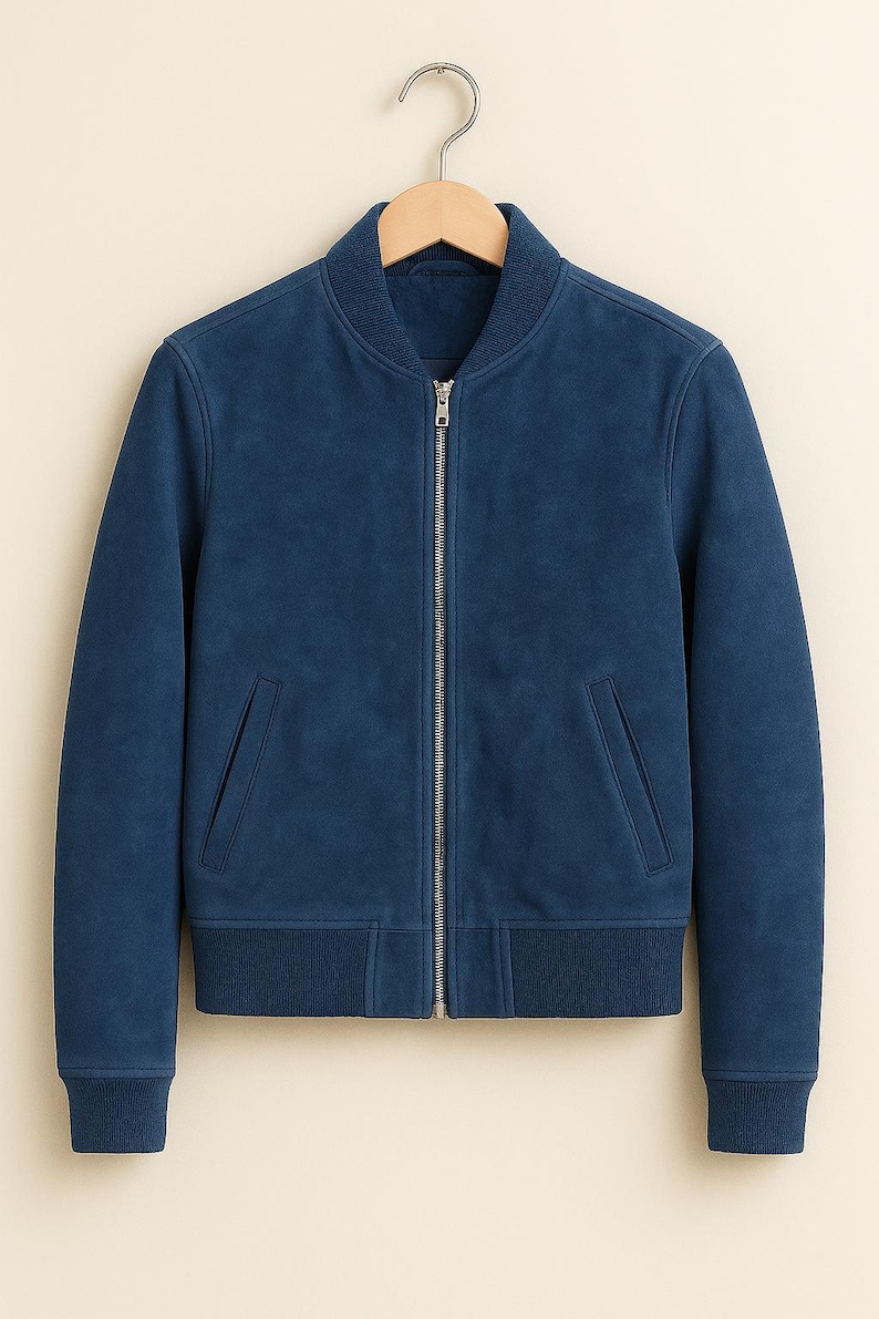 May include: A blue suede bomber jacket with a silver zip. The jacket features two front pockets and ribbed cuffs and hem. It is hanging on a wooden hanger.