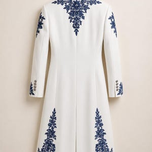 May include: A long white coat featuring intricate dark blue floral embroidery on the collar, shoulders, and lower panels. The coat has long sleeves with four buttons at each cuff and a central back vent, hanging on a wooden hanger.