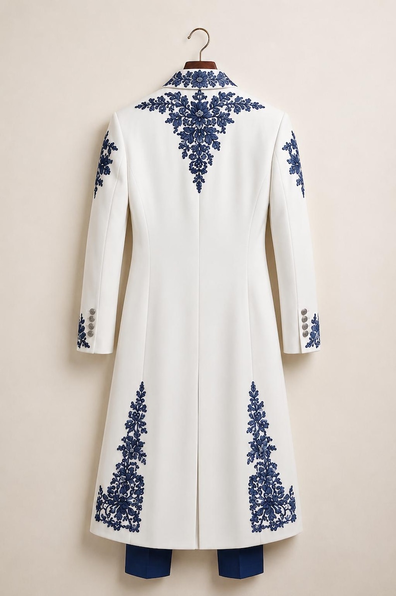 May include: A long white coat featuring intricate dark blue floral embroidery on the collar, shoulders, and lower panels. The coat has long sleeves with four buttons at each cuff and a central back vent, hanging on a wooden hanger.