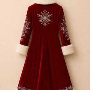May include: A long, deep red velvet coat featuring white faux fur trim on the collar and cuffs. The coat is decorated with intricate white snowflake embroidery on the back, sleeves, and along the bottom hemline, hanging on a wire hanger.