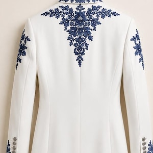 May include: A white blazer featuring intricate dark blue floral embroidery across the upper back, collar, and shoulders. The long-sleeved garment includes four small buttons on each cuff and a structured, tailored fit with a center back seam.