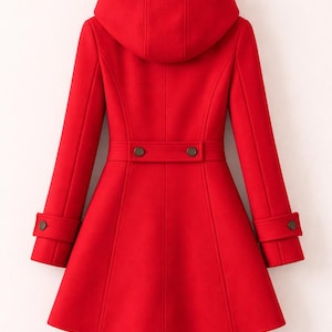 May include: A bright red hooded coat featuring a flared silhouette, long sleeves with buttoned cuffs, and a horizontal back waist tab secured with two dark buttons. The coat is displayed on a wooden hanger against a plain background.