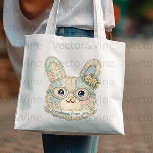 Könnte beinhalten: Weiße Tragetasche mit einem skurrilen Hasenmuster. Der Hase hat ein Patchwork-Design, trägt eine Brille und einen Blumenkranz. Der Text "Somebunny loves you" steht darunter. Die Tasche wird von einer Person gehalten.