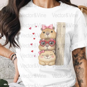 May include: White t-shirt featuring a stack of three cartoon groundhogs. The top and bottom groundhogs have pink bows, while the middle one wears heart-shaped glasses. Small red hearts are scattered around the groundhogs. The text "Vector&Vine" is printed on the shirt.