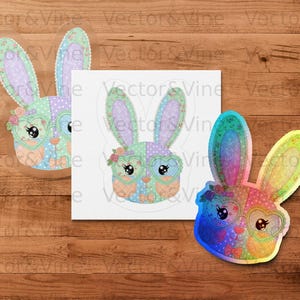 Può includere: Tre disegni colorati di testa di coniglio con occhiali a forma di cuore e accenti floreali. Uno è un adesivo, uno è una stampa su carta bianca e l'altro è un adesivo olografico. Lo sfondo è una superficie di legno.