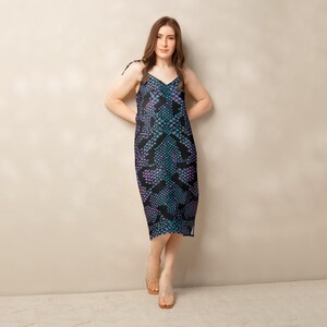 May include: A midi-length slip dress with a snake-skin pattern in teal, purple, and black. The dress has thin straps tied at the shoulders and a V-neckline. The model is wearing clear sandals.
