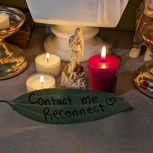 May include: A green leaf with the black handwritten text Contact me Reconnect and a heart symbol. Behind the leaf are two small white tea light candles, a red pillar candle, a small figurine of a person, and a large white bust sculpture.