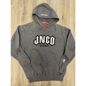 May include: A heather gray hooded sweatshirt featuring the white and black embroidered text JNCO LOS ANGELES on the chest. The hood displays JNCO JEANS text, a small red tag is on the sleeve cuff, and a white skull and crossbones patch is on the arm.