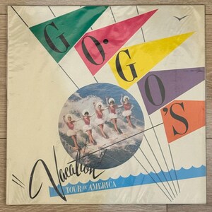 May include: Album cover for The Go-Go's "Vacation" Tour of America. The cover features the band's name in large, colorful flags. A circular image shows the band members in red and white outfits water skiing. The word "Vacation" is in cursive.