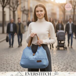 May include: A light blue denim hobo bag with tan leather handles and a small brown rectangular patch on the front. The bag features a single button closure at the top center and a structured base. The text EVERSTITCH is visible in the top right corner.