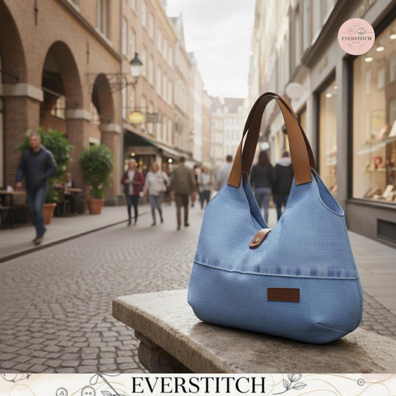 May include: A light blue hobo-style handbag made of a denim-like fabric, featuring two brown leather shoulder straps, a central snap button closure, and a small rectangular brown leather brand label on the lower front section of the bag.