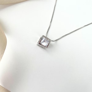 Puede incluir: Un collar plateado con un colgante cuadrado que contiene una gema transparente de talla cuadrada. El colgante cuelga de una cadena fina. El collar se presenta sobre una superficie blanca, con una cinta color crema.