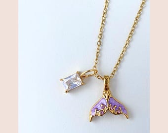 May include: Gold-toned necklace featuring a purple mermaid tail pendant with intricate detailing and a rectangular clear crystal charm. The chain is delicate, and the design is set against a white background.
