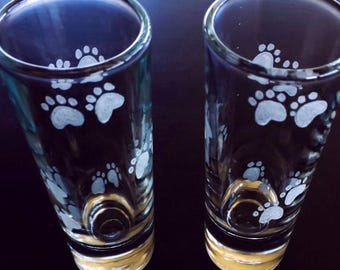 Puede incluir: Dos vasos de chupito transparentes con un diseño de huellas de patas. Las huellas son blancas y están grabadas en el cristal. La base de los vasos tiene una banda dorada.