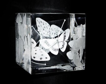 Könnte beinhalten: Ein klarer, quadratischer Glasblock mit geätzten Schmetterlings- und Blumendesigns. Die Schmetterlinge haben detaillierte Flügelmuster, und die Blumen haben zarte Blütenblattformationen. Der Block ist vor einem schwarzen Hintergrund platziert.