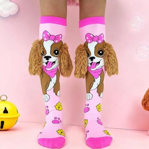 May include: A pair of pink knee-high socks featuring a cartoon dog design. The socks have a pink bow, brown ears, and a pink bandana. The lower part of the socks have paw prints and yellow bells. The socks are worn by a person.