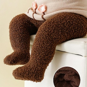 May include: Brown, fuzzy baby leggings with attached feet. The leggings are paired with a beige sweater with floral accents. The image includes a close-up showing the inside of the leggings, with the text "keep warming".