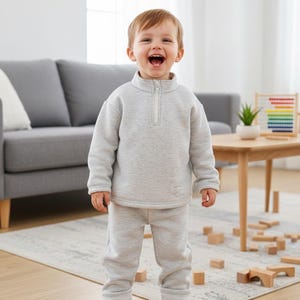 May include: A toddler wearing a light gray sweatshirt with a quarter-zip neckline and matching sweatpants. The child is also wearing white sneakers with velcro straps. The background includes a gray sofa, a wooden table, and a rug.