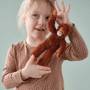 May include: A plush, brown orangutan toy with a smiling face and outstretched arms. The stuffed animal is held up by a child. The toy has a soft, furry texture and is designed to resemble a real orangutan.