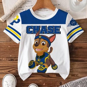 May include: White t-shirt with blue and yellow accents, featuring the word "CHASE" and a cartoon dog character. The shirt has short sleeves with a basketball graphic and the number "2" on the sleeve.