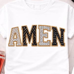 Pode incluir: Camiseta branca com a palavra "AMEN" em um design de joias. As letras são contornadas em ouro e preenchidas com pedras preciosas claras e pretas. A camiseta é de estilo casual.
