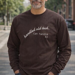 以下が含まれることがあります： 「handled old tech.」と「Can handle AI」という白い文字が入った、長袖の茶色のシャツ。 シャツは無地で、柔らかい素材でできているようです。