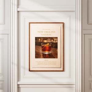 May include: A framed print featuring a cocktail, the New Orleans Sazerac, with a glass of amber liquid and a lemon peel garnish. The print is in a wooden frame and mounted on a cream-colored background. The text "New Orleans Sazerac" is visible.