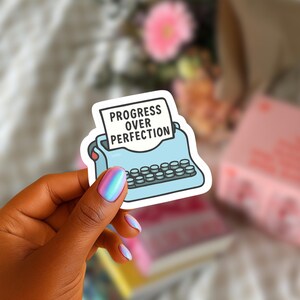 May include: A sticker featuring a light blue typewriter with the words "PROGRESS OVER PERFECTION" on a white paper. The sticker is held by a hand with iridescent nail polish.