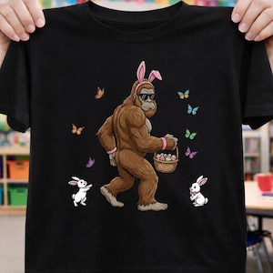 May include: Black t-shirt featuring a cartoon Bigfoot wearing sunglasses and bunny ears, carrying a basket of Easter eggs. The design includes butterflies and two white rabbits. A fun, whimsical design for Easter.