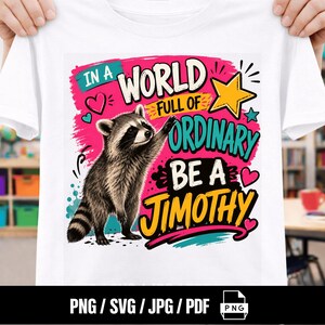 Könnte beinhalten: Ein weißes T-Shirt mit einem Grafikdruck eines Waschbären und dem Text IN A WORLD FULL OF ORDINARY BE A JIMOTHY. Das Design enthält einen gelben Stern, rosa und türkisfarbene Herzen sowie farbige Pinselstrich-Akzente in Pink, Türkis und Gelb.