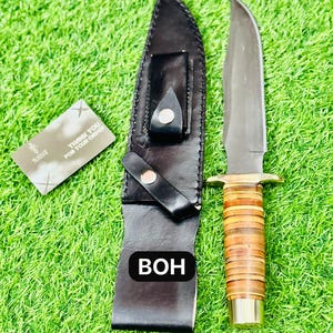 May include: A hunting knife with a leather sheath. The knife has a silver blade, a brass guard, and a handle wrapped in brown leather. The sheath is black leather. A small card with text is also present.