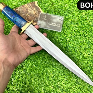 Può includere: Un pugnale con una lama argentata lucida e un manico con dettagli blu e oro. L'impugnatura ha una ghiera color oro e un motivo blu venato legno. La lama è tenuta in una mano su uno sfondo verde erboso. Il testo "BOH" è nell'angolo in alto a destra.