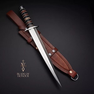 Può includere: Un pugnale con una lama in acciaio lucido e un'impugnatura con fasce alternate nere, marroni e beige. Il pugnale è riposto in un fodero di cuoio marrone con un passante per cintura. Il testo "BLADE OF HONOUR" è visibile.